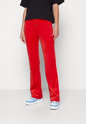 Osoba nosząca jasnoczerwone spodnie o prostych nogawkach z troczkiem w pasie, czarny top z krótkim rękawem oraz białe i niebieskie trampki, stojąca na białym tle.