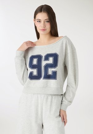 Giovane donna con lunghi capelli castani indossa una felpa grigio chiaro con scollo ampio e grande numero 92 blu scuro, abbinata a pantaloni coordinati.