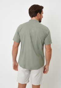 Kortærmet skjorte i lys olivengrøn, lavet af blødt stof, med button-down krave og buet forkant. Båret med lyse beige shorts.