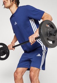 Tenue de sport navy avec des bandes blanches sur les côtés ; manches courtes ; shorts. Le modèle soulève un poids, mettant en valeur la texture du tissu et le logo de la marque.