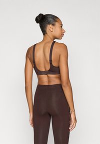 Hnedá športová podprsenka s nastaviteľnými ramienkami, hladkým materiálom a háčikovým zapínaním vzadu, doplnená o hnedé legíny v rovnakom odtieni.