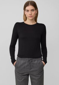 Eine Frau steht mit Händen in den Taschen ihrer grauen Kordelhosen. Sie trägt ein enges schwarzes Langarmoberteil vor einem schlichten grauen Hintergrund.