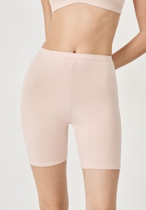 Frau, die hoch taillierte, hellbeige, enganliegende Biker-Shorts und passenden Sport-BH trägt, vor einem schlichten hellen Hintergrund stehend.