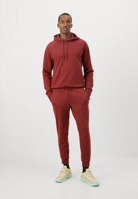 Muž v burgundské mikině a odpovídajících joggerech s béžovými teniskami, stojící s rukama v kapsách na bílém pozadí.