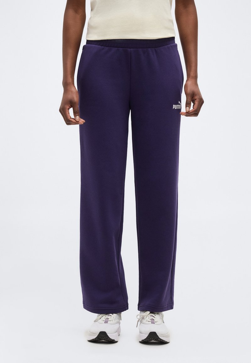 Άτομο με σκούρο μοβ φόρμας Puma, άσπρα αθλητικά παπούτσια και ανοιχτό μπεζ αμάνικο τοπ, στέκεται μπροστά σε απλό φόντο.