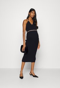Vestido preto com nervuras, decote em V, design sem mangas e cinto de corrente dourada. Combinado com sapatos de salão pretos com bico fino e uma clutch preta peluda.