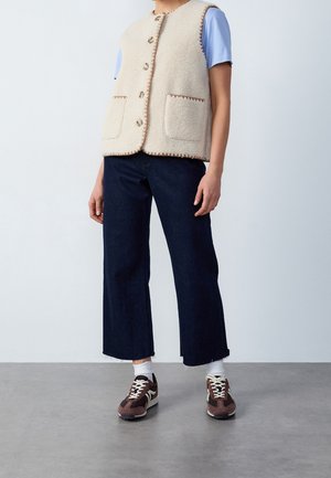 Persona che indossa una camicia azzurro chiaro a maniche corte, un gilet in pile crema con bottoni e tasche, pantaloni corti scuri a gamba larga, calzini bianchi e scarpe da ginnastica marroni.