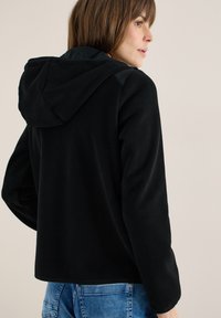 Femme de profil portant une veste polaire noire à capuche avec manches longues et un jean bleu, sur un fond uni.