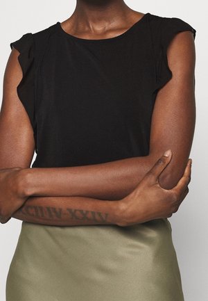Personne aux bras croisés portant un haut sans manches noir et une jupe vert olive, montrant un tatouage de chiffres romains sur l'avant-bras gauche.