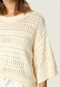 Krämfärgad stickad tröja med lös passform, vida ärmar och texturerade mönster med öppna nätsektioner, vilket framhäver en mysig design.