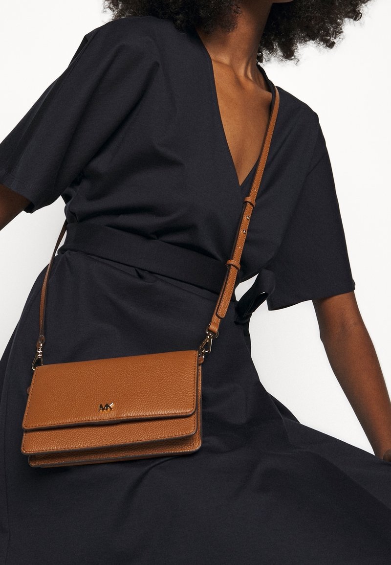Femme portant une robe noire à col en V et un sac bandoulière en cuir marron avec une sangle réglable et des ferrures dorées.