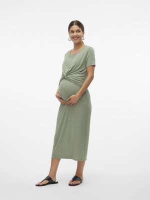 Rase naine seisab, naeratab, kannab rohelist soonikkoes lühikeste varrukatega kleiti sõlmedetailiga ja musti liugjalanõusid valge tausta ees.