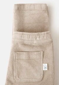 Pantaloni bej din corduroy pliați, având un buzunar la spate și un mic etichetă albă cu un logo de elefant. Detalii cu textură în relief vizibile.