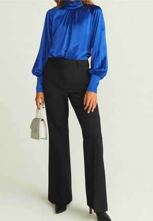 Kvinde iført en skinnende blå bluse med lange ærmer og sorte skræddersyede bukser, holder en lille sølvfarvet håndtaske og har sorte spidse sko på.