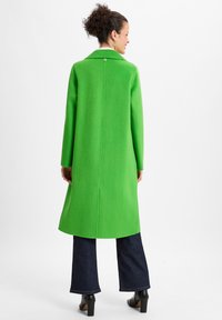 Green wool coat with a notched collar, back vent, and button detail, paired with dark denim jeans and black heeled boots.
