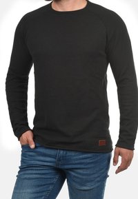 Chemise noire à manches longues avec un col rond et des manches raglan. Présente un tissu texturé et une petite étiquette en cuir marron près de l'ourlet.