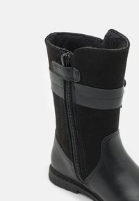 Musta nilkkuri, joka on valmistettu teksturoidusta mokasta ja sileästä nahasta. Varustettu vetoketjulla ja erottuvilla vaakasuorilla hihnoilla lisäyksinä.