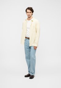 Ανοιχτό κίτρινο λινό πουκάμισο πάνω από λευκό μπλουζάκι, μπλε τζιν και σκούρα καφέ παπούτσια· το μοντέλο στέκεται σε ουδέτερο φόντο.