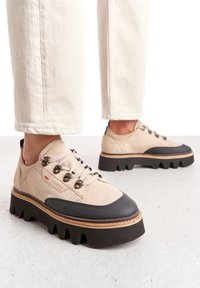 Scarpe in suede beige con punta in gomma nera, suola spessa e seghettata, e ganci metallici per i lacci. Accentuate da una striscia in pelle marrone lungo la suola.