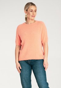 Krótki rękaw, koralowy sweter z dzianiny, okrągły dekolt i miękka tekstura, w połączeniu z ciemnoniebieskimi dżinsami. Posiada luźny krój i podwinięte rękawy.