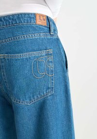 Jeans de mezclilla azul con una etiqueta de cuero en la cintura trasera y un único bolsillo trasero que presenta un detalle de logo bordado.