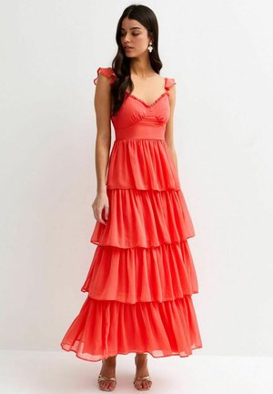 Donna con capelli lunghi e scuri che indossa un abito maxi corallo a balze con spalline arricciate, in piedi e che guarda a sinistra contro uno sfondo semplice.