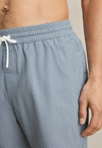 Modré šortky so vzorom, s elastickým pásom a bielou šnúrkou. Materiál má jemnú textúru a obsahuje geometrický dizajn v tvare diamantu.