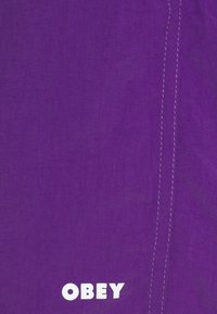 Lila tyg med en framträdande vit "OBEY"-logotyp och kontrasterande sömdetaljer, som visar en mjuk yta och en enfärgad design.