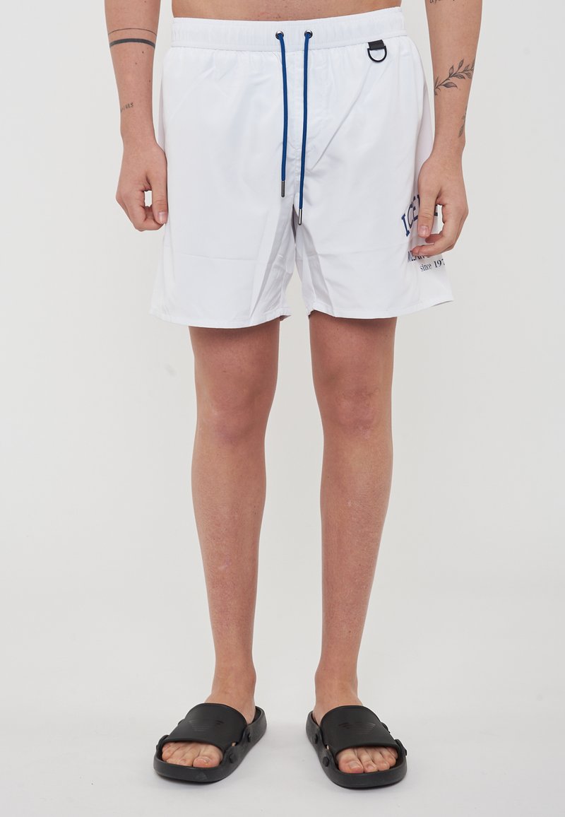 Shorts de bain blancs avec un cordon de serrage, des accents bleus et des poches latérales. Associés à des sandales noires à enfiler, avec une surface texturée.