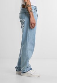 Lys blå denim jeans med en avslappet passform, med slitte detaljer og en lapp på baklommen. Brukt med hvite sneakers.