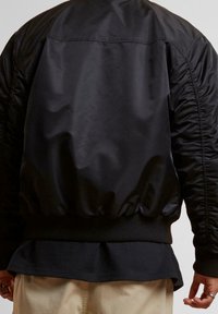 Veste bomber noire avec un extérieur lisse, ourlet côtelé et détails froncés sur les manches. Portée sur une chemise sombre texturée.