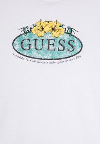 Valkoisesta puuvillapaidasta löytyvä kulunut turkoosi soikea logo, jossa lukee "GUESS" paksulla mustalla tekstillä, ja ylhäällä on kaksi keltaista hibiskuskukkaa.