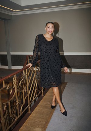 Une femme pose près de la rampe d’un escalier, vêtue d’une robe noire transparente à pois et de chaussures noires à talons.