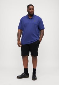 Námornícky modré polo tričko s golierom a žltým logom, skombinované s čiernymi krátkymi nohavicami. Čierne topánky a ponožky dopĺňajú tento outfit.