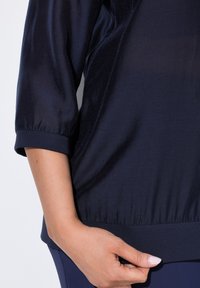 Tummansininen toppi, joka on valmistettu kiiltävästä ja sileästä kankaasta, jossa on kolme neljästä hihoista ja laskostettu helma. Ominaisuuksiin kuuluu pehmeä tekstuuri ja kumitettu hihansuu.