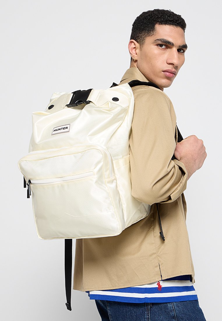 Joven con chaqueta beige y camisa a rayas azules, con una mochila crema de Hunter al hombro, mirando por encima del hombro.