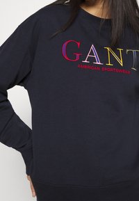 Felpa blu navy con testo ricamato colorato "GANT AMERICAN SPORTSWEAR" in rosso, rosa e giallo. Texture del tessuto morbida, vestibilità rilassata.