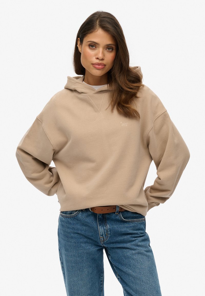 Sweatshirt beige à capuche avec des manches larges et une poche kangourou, arborant un petit logo sur la poitrine. Coupe décontractée associée à un jean bleu.