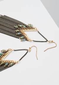 Gold- und schwarze Kettenohrringe mit grünen Perlenakzenten; elongated, geschichtetes Fransen-Design; leicht mit Hakenverschlüssen.