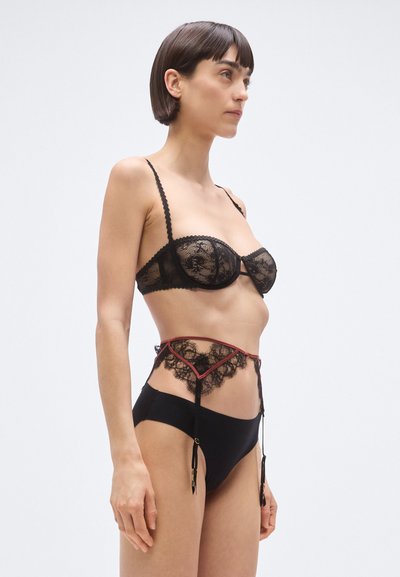 Čierna čipková podprsenka s vlnitými okrajmi, skombinovaná s čiernymi nohavičkami s vysokým pásom, ktoré majú čipkový lem a červené akcenty. Súčasťou sú nastaviteľné ramienka.