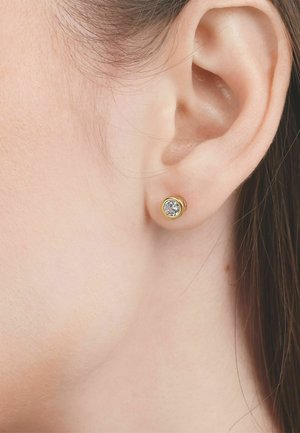 Close-up van het oor van een vrouw met een kleine, ronde gouden oorbel met een heldere edelsteen in het midden.