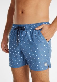 Shorts de bain bleu en tissu léger, avec de petits motifs de feuilles blanches, une taille élastique et des poches latérales.
