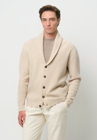 Beige gestrickte Strickjacke mit einem Schalkragen, ausgestattet mit fünf dunklen Knöpfen und strukturiertem Rippenmuster, kombiniert mit einem hellgrauen Hemd und cremefarbenen Hosen.