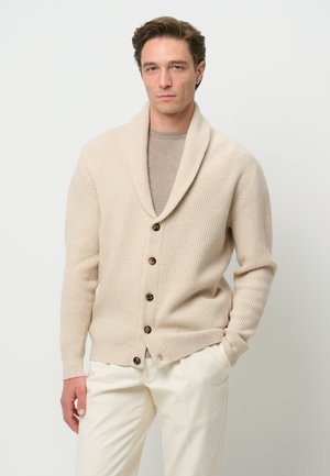 Beige knitted cardigan with a shawl collar, featuring five dark buttons and ribbed texture, paired with a light gray shirt and cream pants.