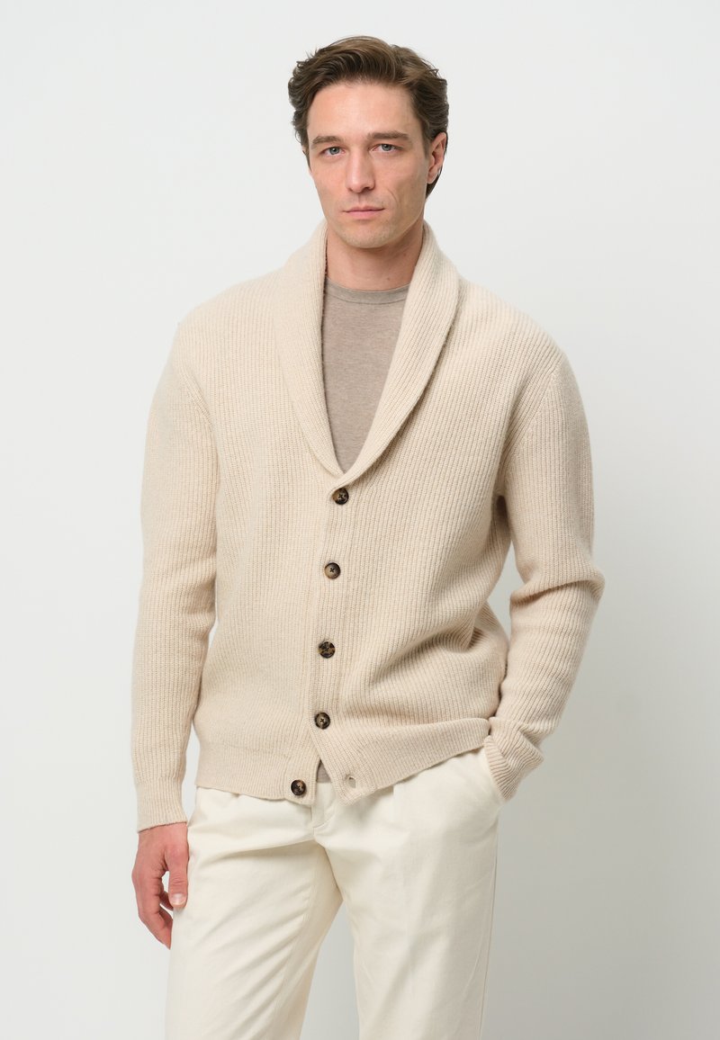 Beige gestrickte Strickjacke mit einem Schalkragen, ausgestattet mit fünf dunklen Knöpfen und strukturiertem Rippenmuster, kombiniert mit einem hellgrauen Hemd und cremefarbenen Hosen.