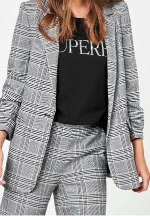 Žena oblečená v šedém kostkovaném blejzru a kalhotách, s černým tričkem, na kterém je částečně viditelný bílý nápis "SUPER".