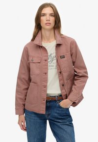 Różowa bawełniana kurtka z kołnierzykiem, dwoma bocznymi kieszeniami i kieszenią na piersi. Nosi się ją na beżowej koszuli i zestawiono z niebieskimi dżinsami.