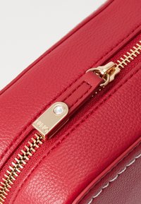 Angolo di una borsa rossa in pelle con una cerniera dorata con un accento in cristallo, superficie testurizzata e dettaglio in bianco lungo il bordo.