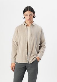 Camisa de botón de pana beige, con cuello, manga larga y bolsillos delanteros, llevada sobre una blusa de color claro, combinada con pantalones grises.