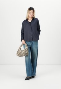 Woman wearing loose dark shirt and wide-leg blue jeans holding a snakeskin-patterned handbag, standing against a plain white background.